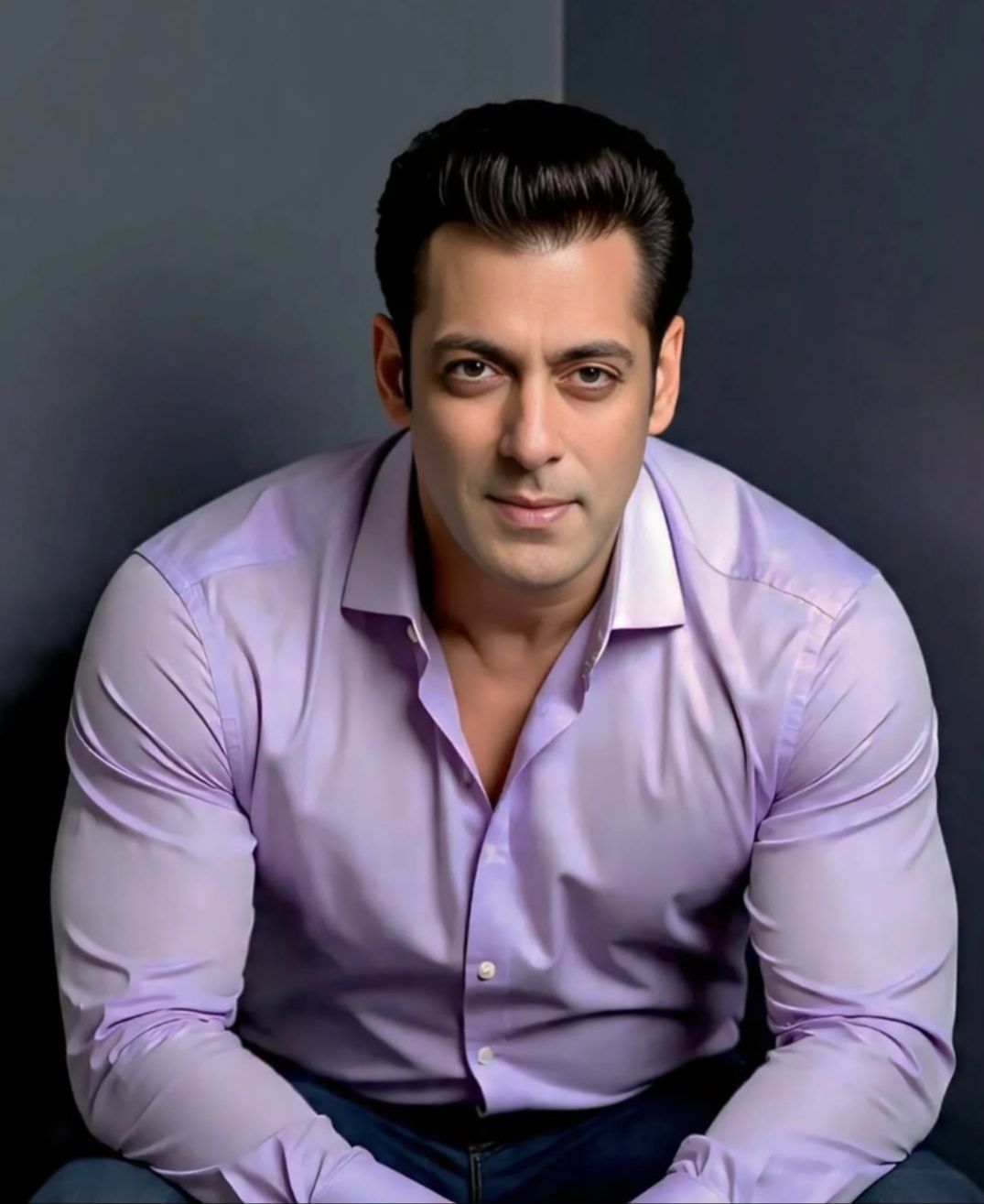 A portrait of Salman Khan seated and leaning forward, wearing a lavender button-down shirt and dark jeans against a dark grey background.