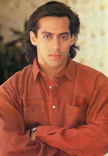 A medium close-up portrait of a young Salman Khan with wavy, neck-length hair, wearing a rust-colored button-down shirt and looking directly at the camera with a calm expression.