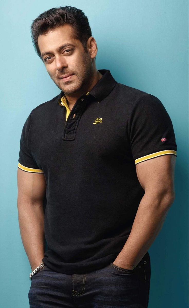 A medium shot of Salman Khan standing against a blue wall, wearing a black Being Human polo shirt and dark jeans with his hands in his pockets.