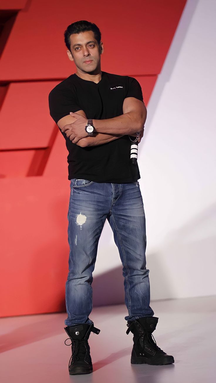 A medium shot of Salman Khan standing against a blue wall, wearing a black Being Human polo shirt and dark jeans with his hands in his pockets.